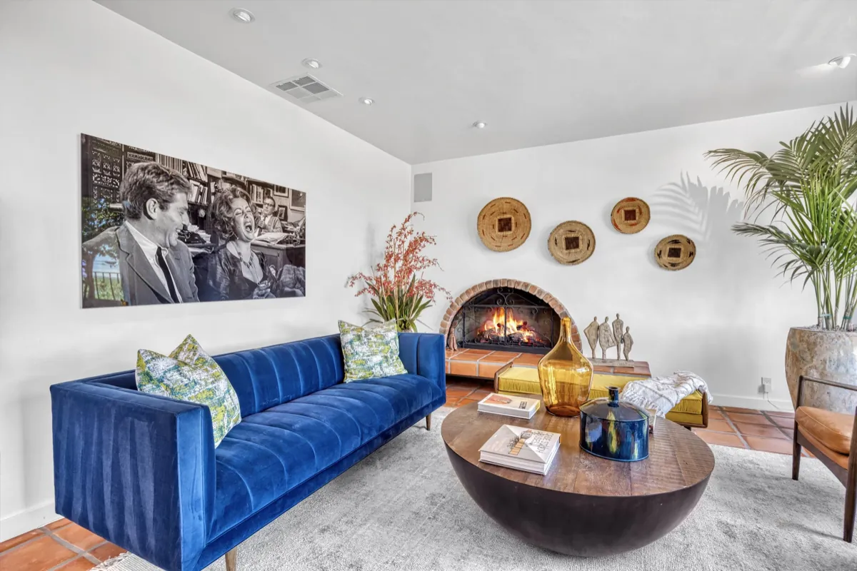 A living area with a blue velvet sofa facing a lit corner fireplace, terracotta tile floor and framed artwork.